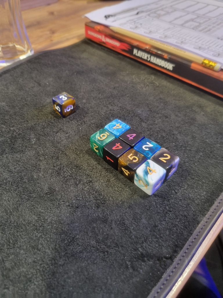 Nine d6 resting on a mat, ready to be rolled for a cast of fireball. In the background can be seen a D&D 5e player's handbook, a pencil, and a character sheet.
